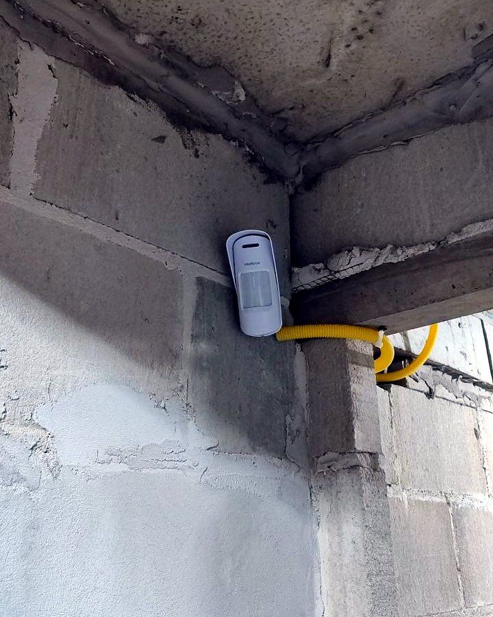 Sensor de presença instalado no canto de uma obra em bloco, com cabo em conduíte amarelo provisório