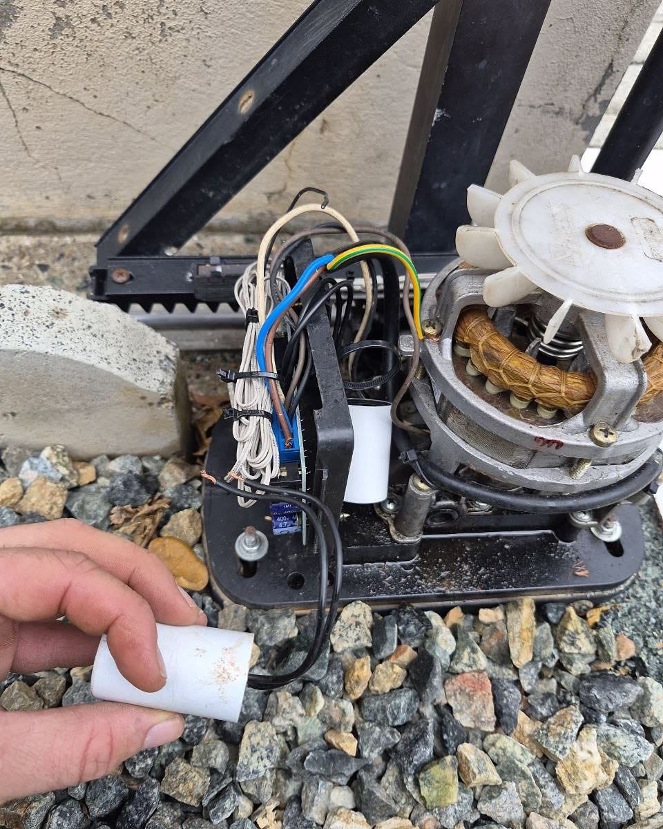 Motor de portão deslizante aberto durante a manutenção, com o técnico segurando o capacitor antigo que foi trocado